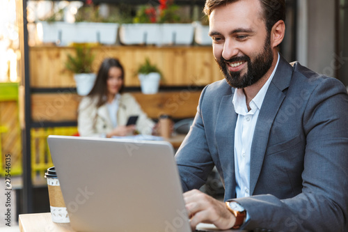 Portrait of masculine businessman smiling and working on laptop while sitting in cafe outdoors