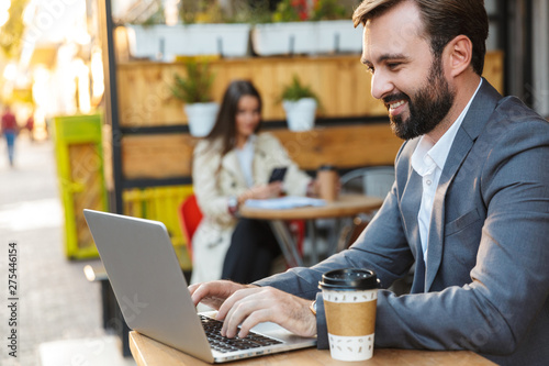 Portrait of handsome businessman smiling and working on laptop while sitting in cafe outdoors