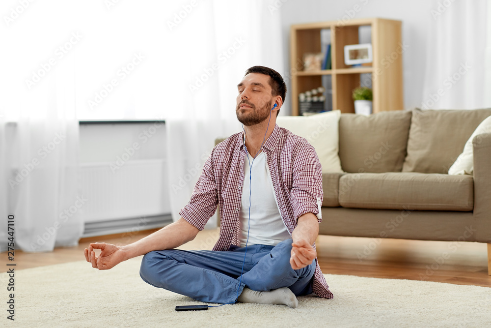 mindfulness, meditation and relaxation concept - man in earphones listening to relaxing music on smartphone and meditating in lotus pose at home
