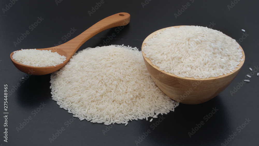 Rice in wooden bowl and heap of rice with spoon on black table