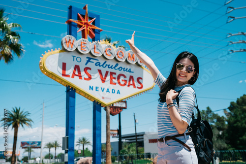 Happy young girl backpacker in sunglasses showing peace gesture and looking at camera while standing by famous billboard in nevada. smiling lady traveler taking picture with las vegas sign on summer
