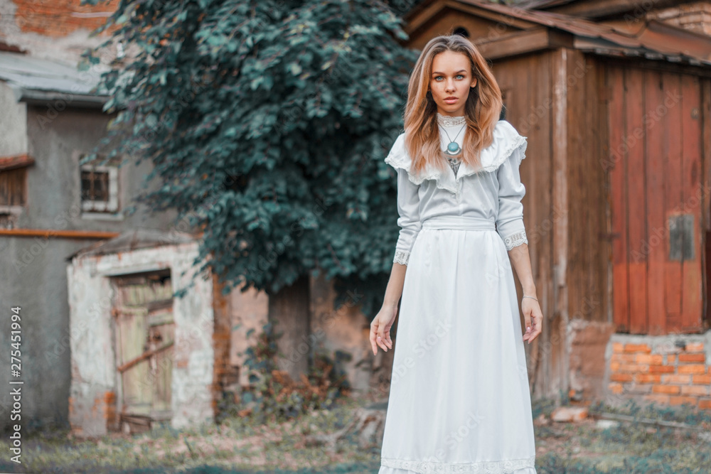 The girl looks straight, stands near the house in a white vintage dress, necklace