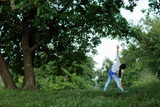 young woman doing triangle pose yoga on yoga mat in white clothes under a big tree in a lush green meadow among dandelions
