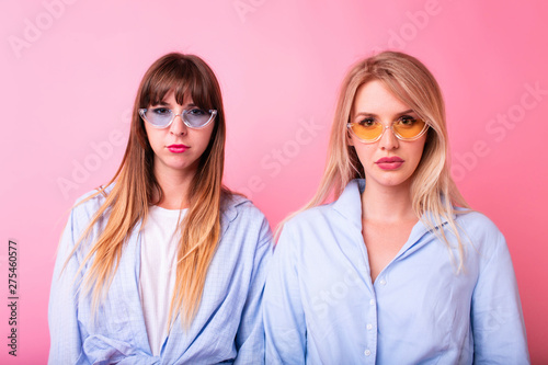 Two pretty upset young smartly dressed girls looking to camera, wearing makeup standing isolated over pink background