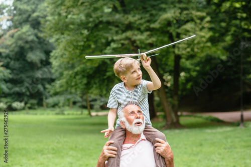 Little boy with airplane model and grandfather raising hands over green park on background enjoying life and nature. Portrait of happy grandfather giving grandson piggyback ride on his shoulders.