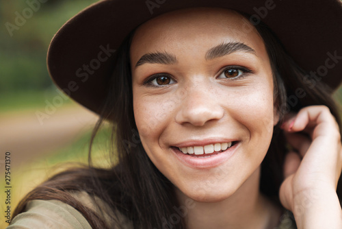 Photo closeup of cheerful pretty woman wearing hat smiling at camera while sitting on grass outdoors