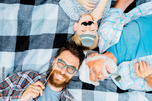 Grandfather,father and son enjoying together lying on a checkered blanket. Tree men of different ages smiling playing with fake mustache, hat, glasses. Top view of boy and his dad, granddad with smile photo