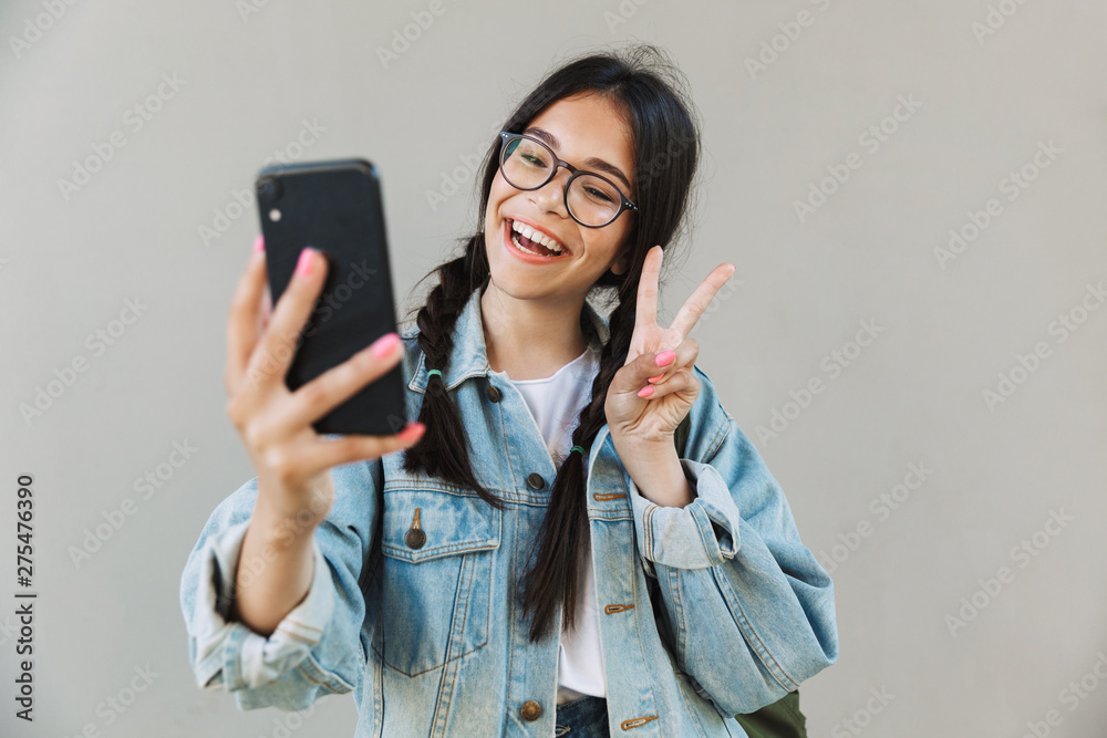 Naklejka Happy cute beautiful girl in denim jacket wearing eyeglasses isolated over gray background using mobile phone take a selfie