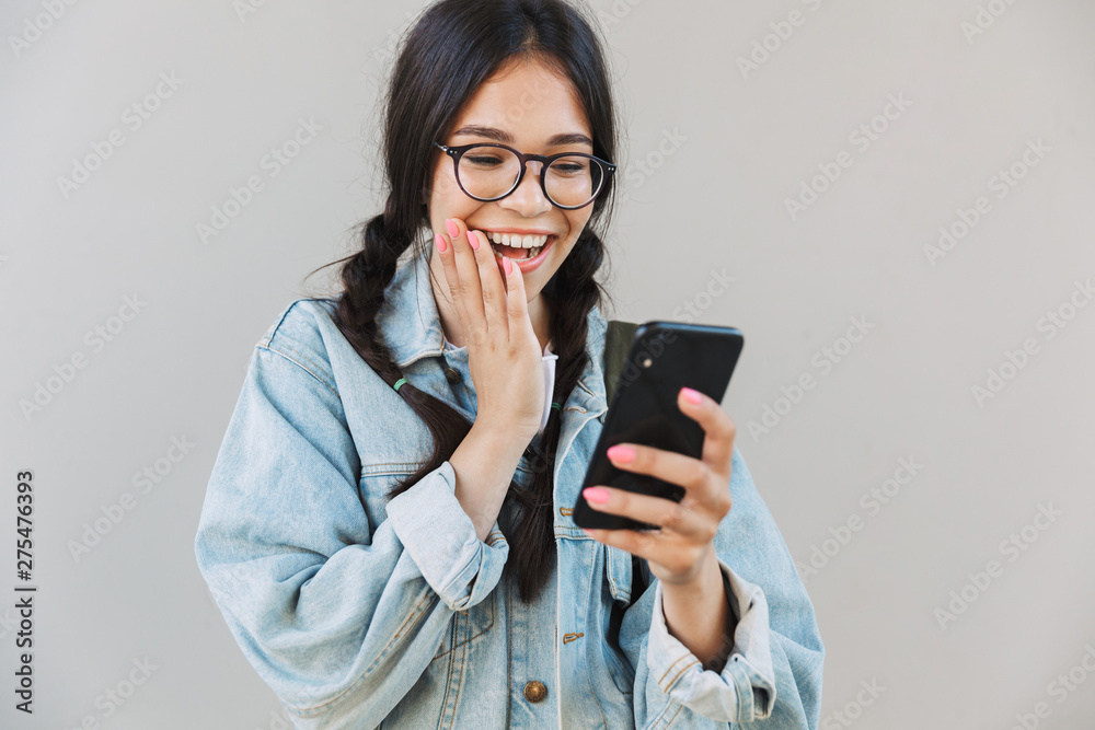 Pleased cute beautiful girl in denim jacket wearing eyeglasses isolated over gray background using mobile phone.