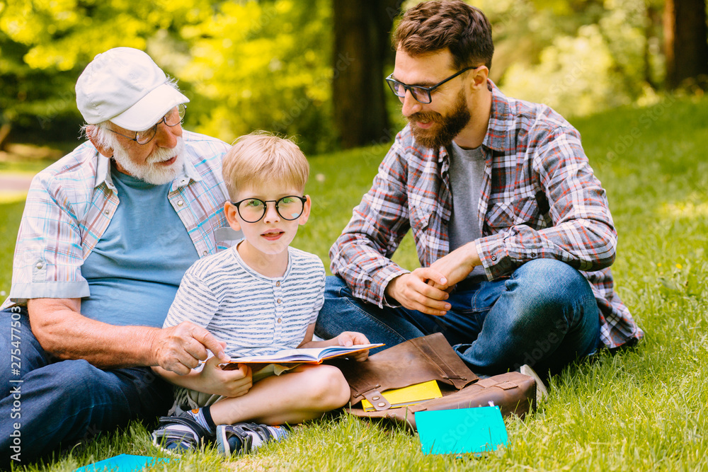 Φωτογραφία happy family of three generation - father, grandfather and blond son sitting on grass at park with books learn to read while getting ready for school
