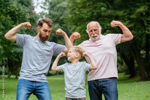 Portrait og happy family - grandpa, father and his son smiling and showing their muscles outdoor in park on background. Three different generation concept. photo