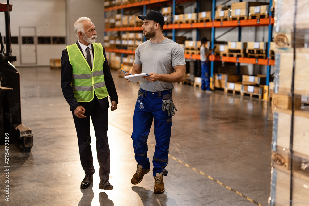 Warehouse worker and mature businessman talking while going through paperwork in industrial building.