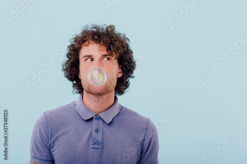 male chewing gum bubbles, curly hair and purple T-shirt, isolated blue background, positive facial emotions, copy space photo