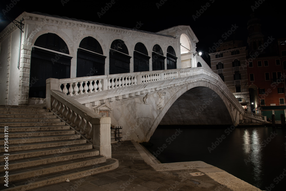 Night photo Rialto Bridge in Venice, Italy. Overview of the white marble bridge illuminated by reflections on the water of the Grand Canal.