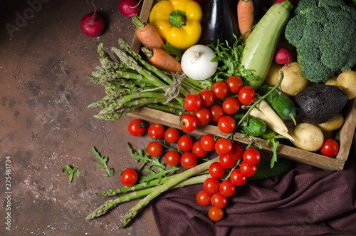 Box with farm vegetables on a dark background. Place for text. Cherry tomatoes, asparagus, broccoli, peppers, eggplants, zucchini, carrots and onions.