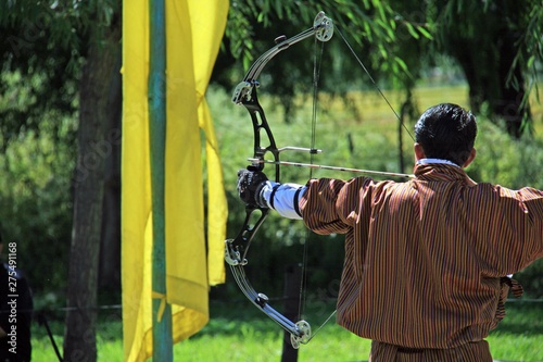 Bogenschütze beim Training im Königreich Bhutan, Himalaya, Asien. Mit modernen Gelenkbogen in traditioneller Kleidung, dem Gho, zielt er im Sonnenschein zum Schuss bereit. Olympia wartet schon.