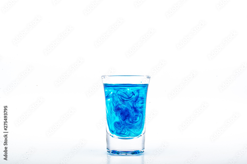 Blue food coloring diffuse in water inside shot glass with empty copyspace area for slogan or advertising text message, over isolated white background. Blue food coloring diffuse in water inside shot glass with empty copyspace area for slogan or advertising text message, over isolated white background.