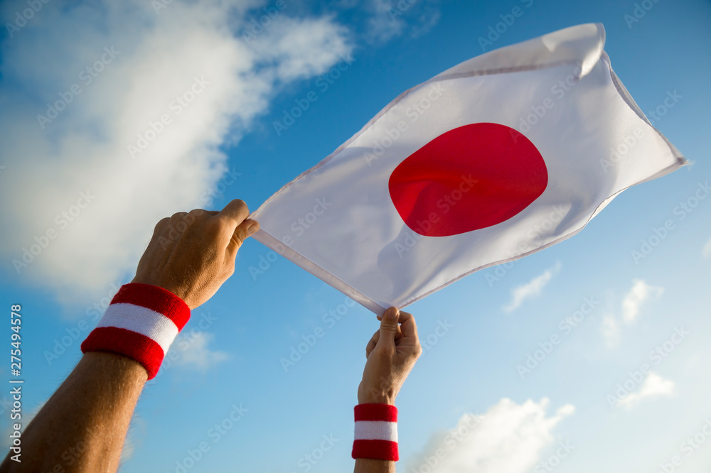 Hands with Japan red and white wristband holding a Japanese flag waving in bright sunny blue sky - obrazy, fototapety, plakaty 