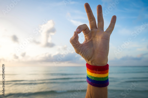 Hand of athlete giving OK sign with gay pride rainbow colors wristband against bright blue sky sea 