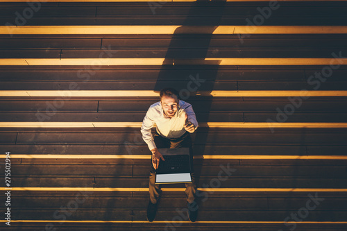 High angle view of businessman sitting on stairs, working on laptop and showing thumbs up at sunset.