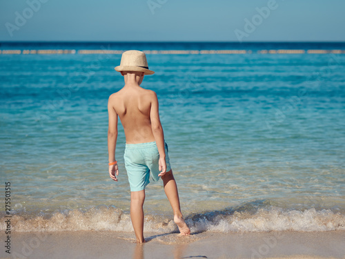 European boy in blue swimming shorts and sunhat is having fun in the sea cost. He is standing on the sand near the ocean and enjoying his summer holidays.