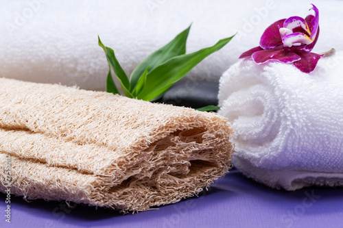 Spa Wellness Concept. Natural Loofah Sponge, rolled up White Towels, stacked Basalt Stones, Bamboo and Orchid Flower on purple background.