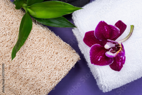 Spa Wellness Concept. Natural Loofah Sponge, rolled up White Towels, stacked Basalt Stones, Bamboo and Orchid Flower on purple background.