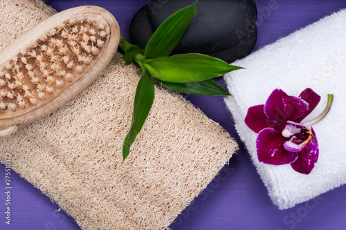 Spa Wellness Concept. Natural Loofah Sponge, Natural bristle Wooden Brush, White Towels, Basalt Stones, Bamboo and Orchid Flower on purple background.