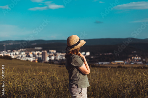 Young woman wearing summer outfit relaxing while looking at cityscape from a high green hill - Casually dressed hipster girl enjoying summer time in nature