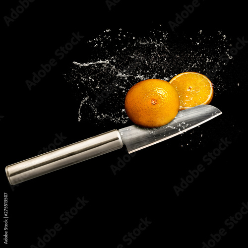 A single metal stainless steel chef knife cutting orange fruit and splashing water, black background