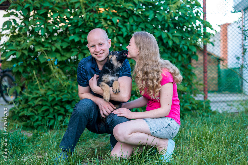 Fototapeta premium Young happy couple with a funny puppy of a German shepherd. A girl and a guy on a green lawn near the house with a new dog. Buying a pet. The joy of first meeting. Copy spase