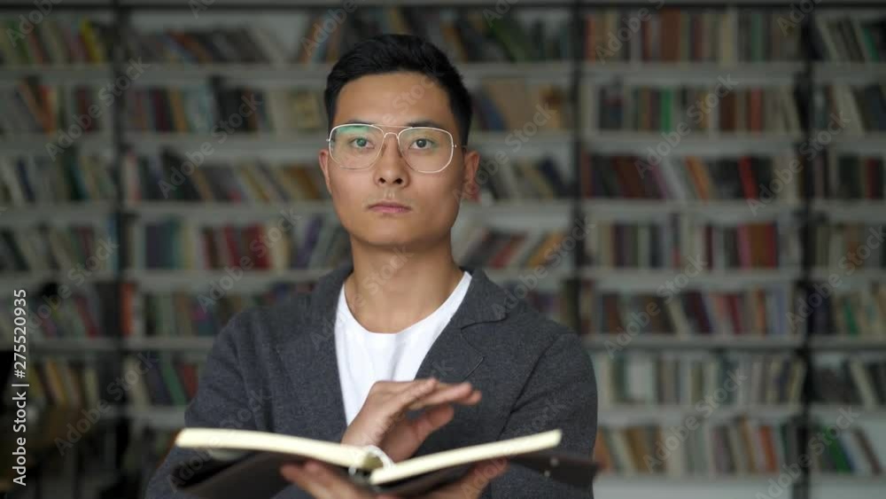 Asian man holding a blocknote swiping in the air in virtual screen with a hand turning over a page on background of bookshelves. Concept of virtual book swiping Asian man holding a blocknote swiping in the air in virtual screen with a hand turning over a page on background of bookshelves. Concept of virtual book swiping