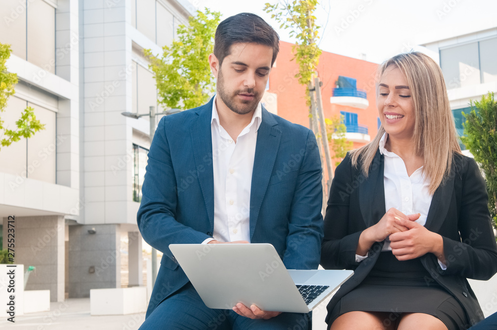 Business man and woman using laptop and working outdoors. Business people wearing formal clothes and sitting with buildings in background. Business work concept. Front view.