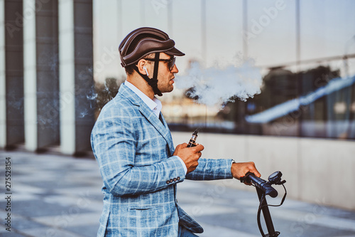 Pensive man in protective helmet and sunglasses is smoking vape while holding his electrical scooter.