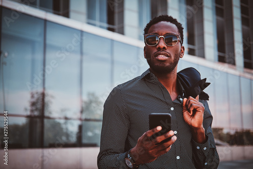Portrait of pensive modern afro man with sunglasses and mobile phone over glass building background.