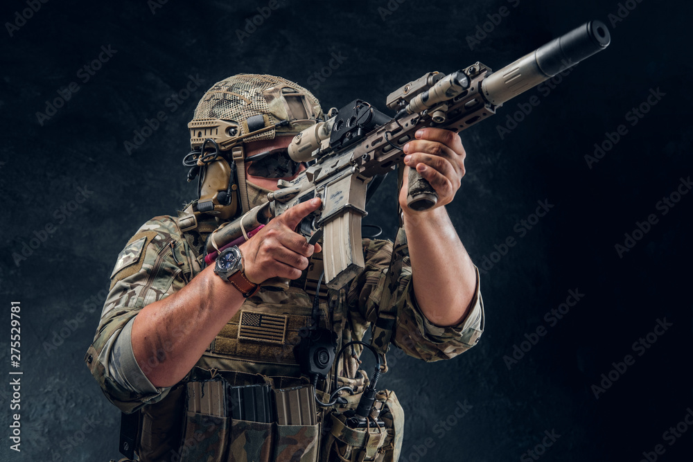 Military man in full equipment with wach on his hand is holding machine gun while posing for photographer over dark background. Military man in full equipment with wach on his hand is holding machine gun while posing for photographer over dark background.