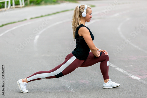 Fitness lifestyle. Young woman in headphones warming up before training doing exercises to stretch her muscles and joints. Workout at the stadium. Healthy life concept © Sergei Shavin