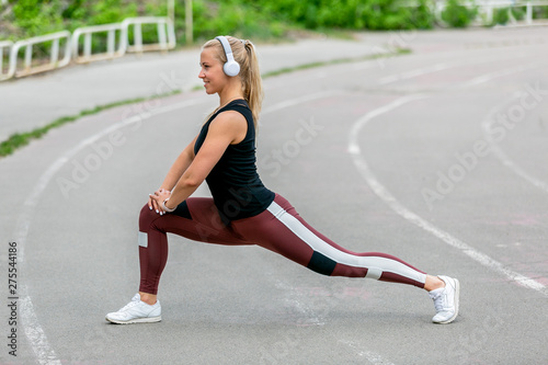 Fitness lifestyle. Young woman in headphones warming up before training doing exercises to stretch her muscles and joints. Workout at the stadium. Healthy life concept