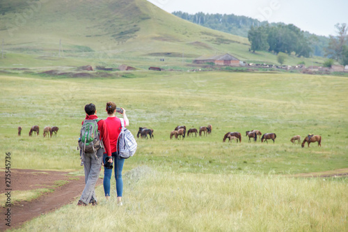 Two young girls with backpacks and photo camera make selfie on phone near rural road. Horse farm pasture with mare and foal. Small village with old houses. Summer landscape with green hills