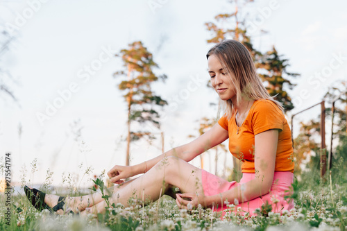Young beautiful blonde girl in a pink skirt and in a coral shirt is sitting on a meadow, looking down and smiling. Beautiful glade with white flowers of clover growing on it. © Svyatoslav Balan