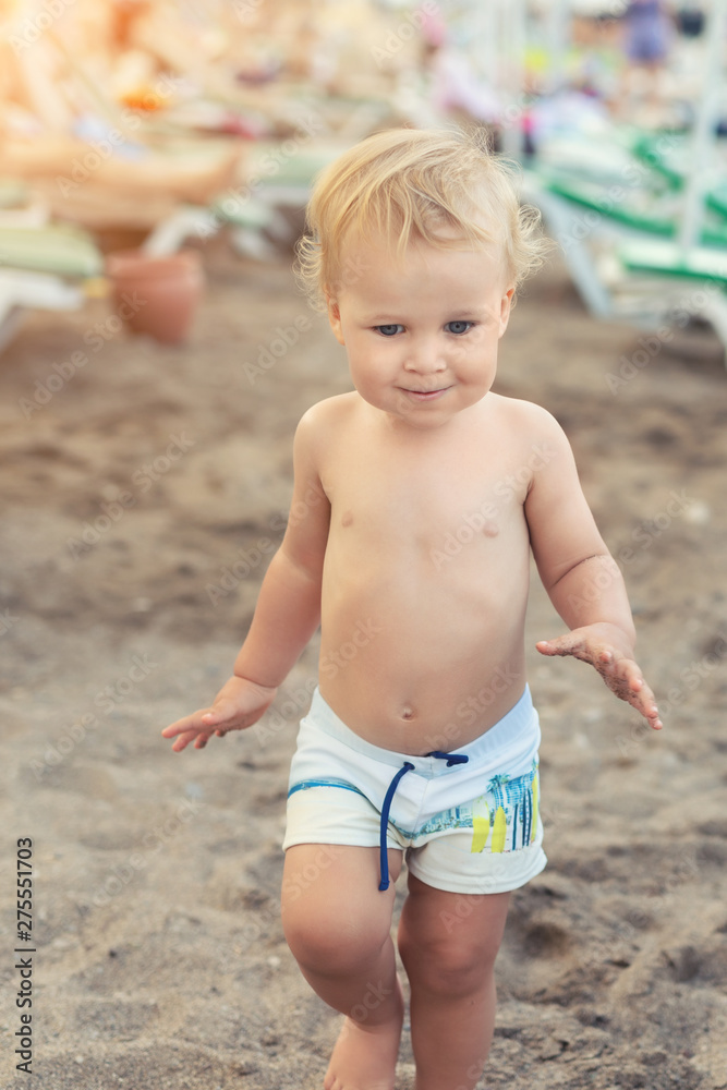Cute caucasian toodler boy walking alone on sandy beach between chaise-lounge. Adorable happy child having fun playing at seaside shore during vacation trip Cute caucasian toodler boy walking alone on sandy beach between chaise-lounge. Adorable happy child having fun playing at seaside shore during vacation trip