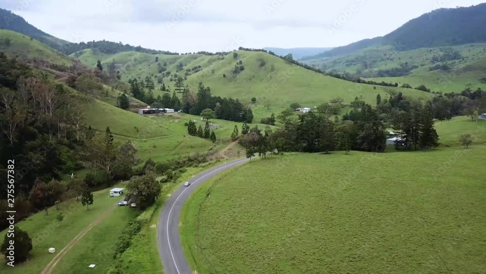 Aerial drone panning above a long winding road through a small rural town in Australia with fresh green grass and tall mountains in the distance on an overcast day Aerial drone panning above a long winding road through a small rural town in Australia with fresh green grass and tall mountains in the distance on an overcast day
