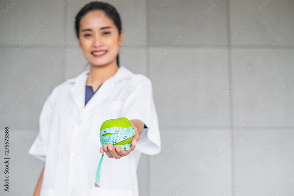 Smiling Asian woman doctor in medical white gown holding a green apple wrap with measuring tape. Health concept. Smiling Asian woman doctor in medical white gown holding a green apple wrap with measuring tape. Health concept.