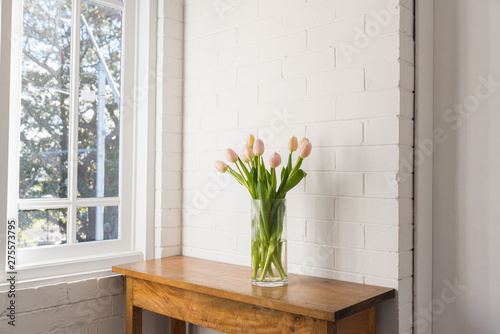 Pale pink tulips in glass vase on wooden sidetable against white painted brick wall and window (selective focus) photo