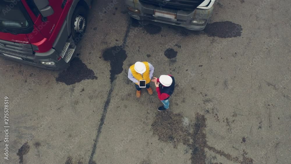 AERIAL. Two workers works on tablet near trucks. Camera zooming out. AERIAL. Two workers works on tablet near trucks. Camera zooming out.