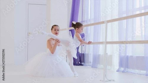 Personal ballet lesson for little girl. Professional teacher is stretchig leg of little girl standing near the barre stand in ballet class, side view. They both are in white tutus and pointes. photo