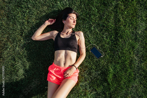 Portrait of a happy young fitness brunette woman lying on the football pitch and listening to music on your smartphone after workout and smiling. Sport in open air and health care concept
