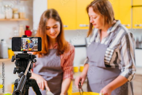 Family culinary video blog. Mother and daughter using smartphone on tripod to film cooking in kitchen. Blur background.