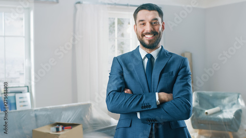 Successful Real Estate Agent in a Suit Smiles and Offers Keys From a New Apartment. Standing in the Middle of Room that Being Renovated. Spacious New House for Sale by Professional Real Estate Broker. photo