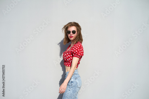 Redhead caucasian girl in red polka-dot pattern blouse and jeans staying near grey background